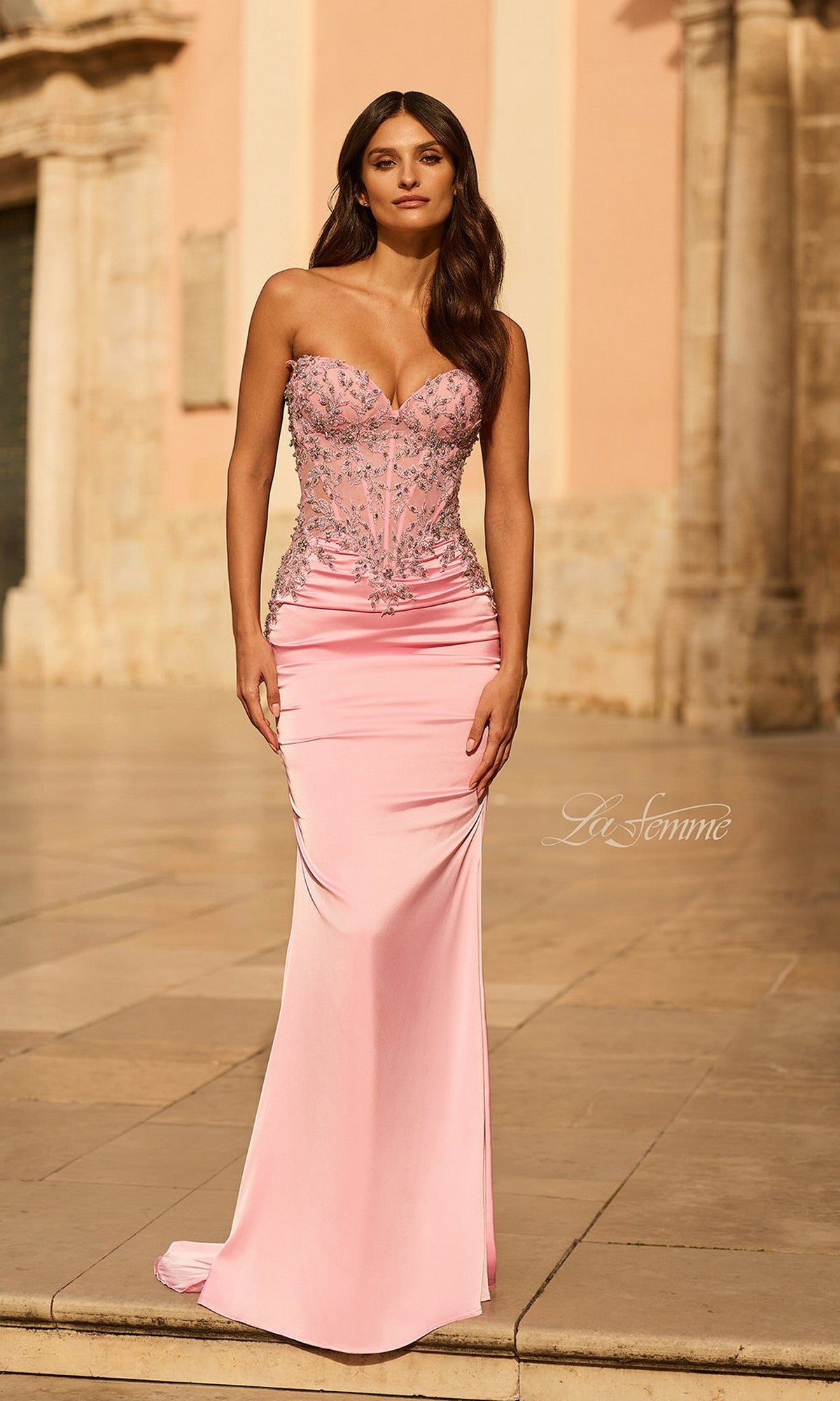 A woman wears the Long Prom Dress: La Femme 33765 in baby pink, featuring a strapless embellished corset bodice with silver details, as she stands outdoors on stone pavement before a historic building.